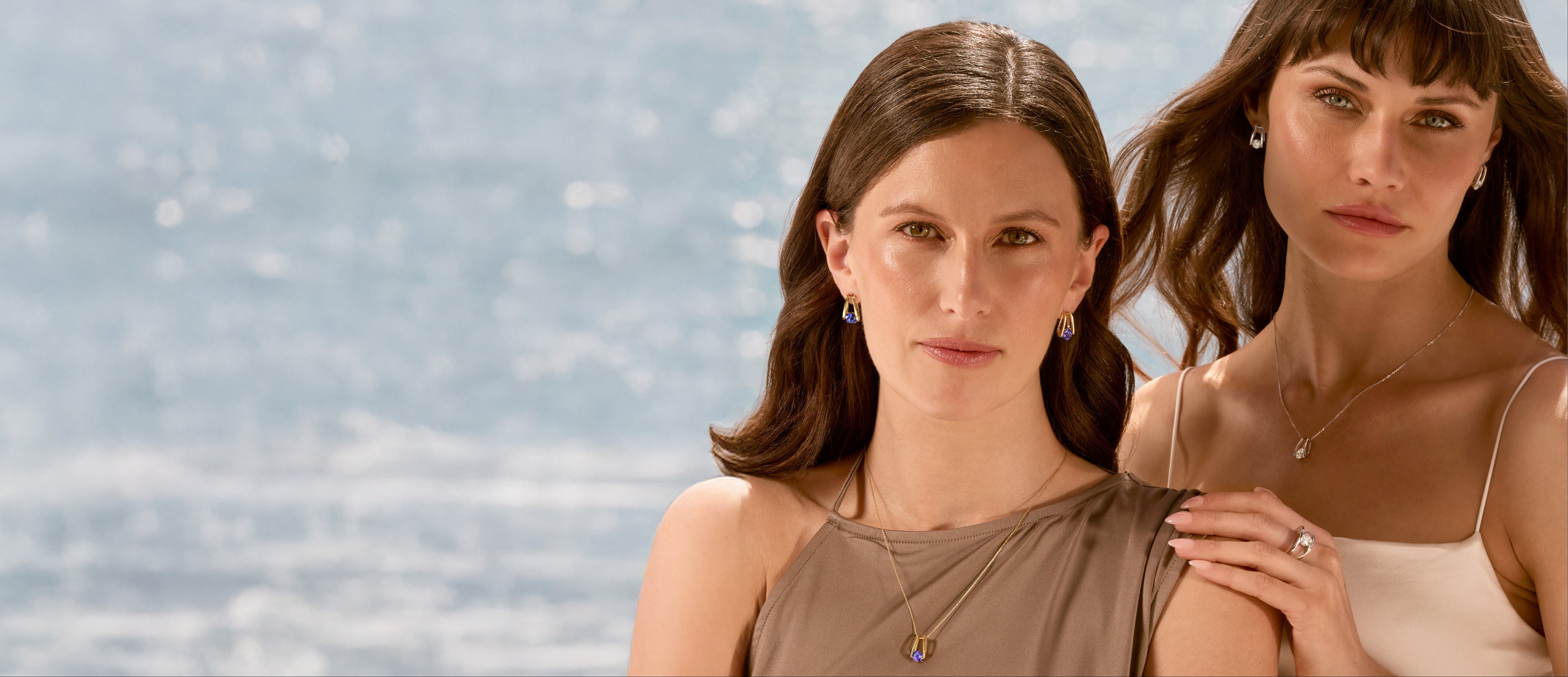 Two women wearing the Shimansky Iconic Millennium Diamond and Tanzanite jewellery collection, standing close together against a light blue water background