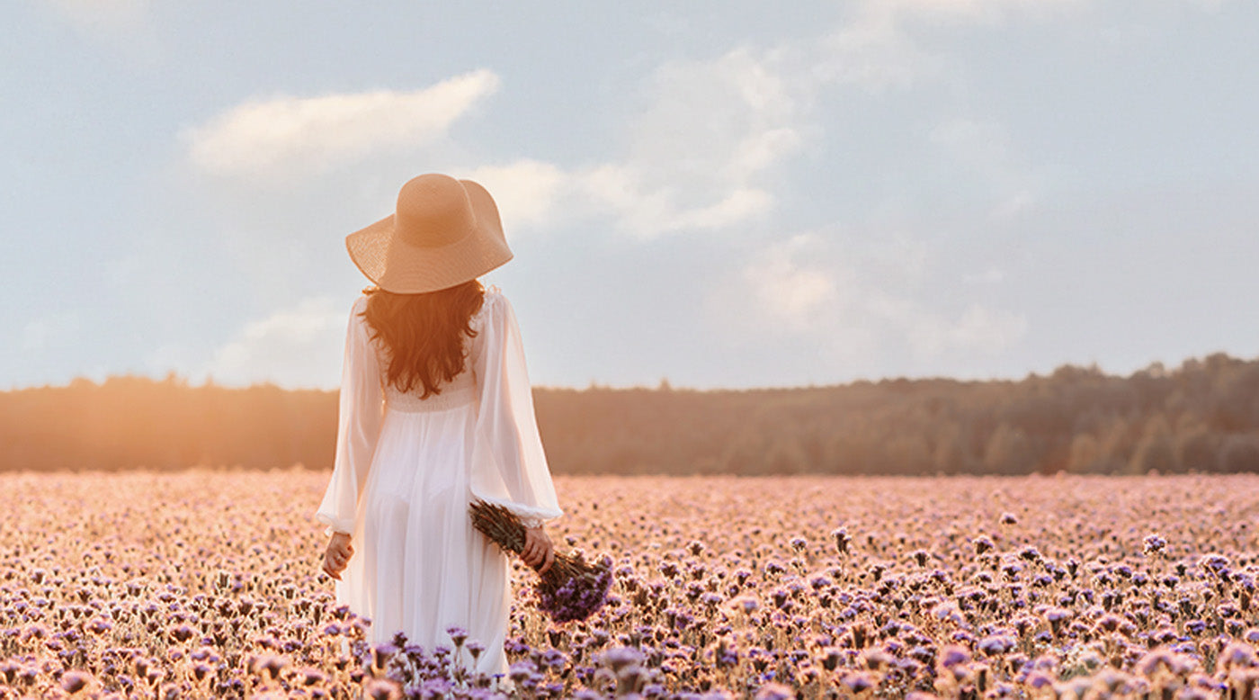 A graceful woman in a white dress and hat strolls through a vibrant field of blooming purple flowers, exuding elegance.