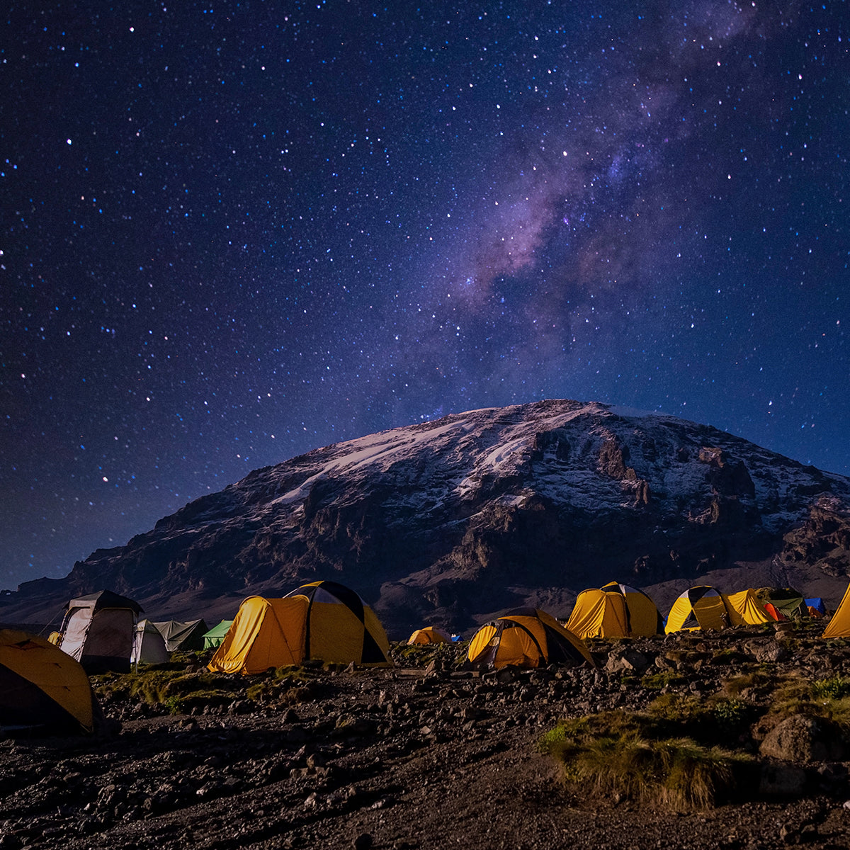 A starry night sky over a mountain campsite, with several yellow and orange tents pitched on rocky terrain. The Milky Way is visible in the background.