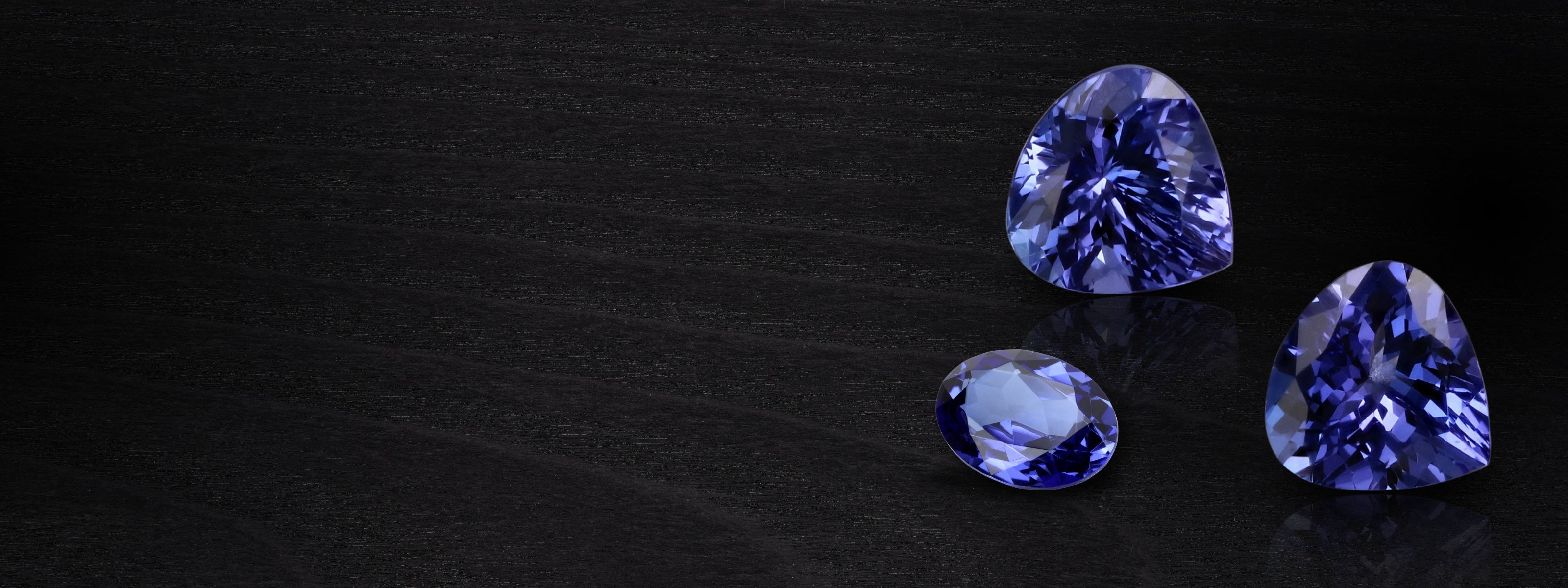 Three exquisite tanzanite gems glisten elegantly on a sleek black surface, showcasing their captivating hues.
